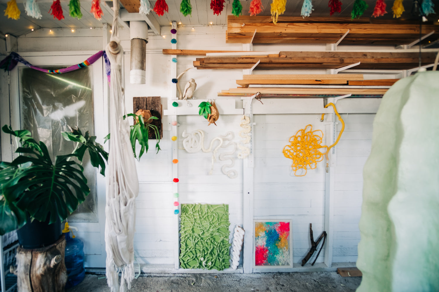 Image of studio wall, with decorative tassels hanging from the ceiling, wood piles, colorful art, plants, and a hammock 